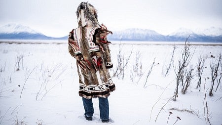 A person wearing an animal fur coat faces away from the camera and looks out at a snowy landscape.