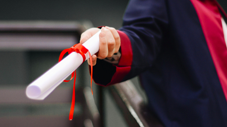 Graduate in cap and gown holding out rolled up diploma.