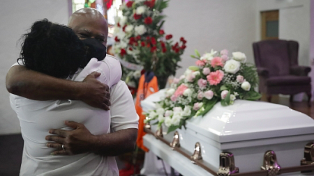 Two people with masks on hug while standing in front of a white coffin.
