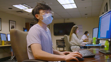A student wearing a mask sits in from of a computer while socially distanced from other students. 