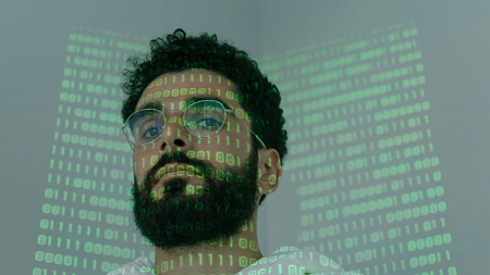 A person with binary code projected onto their face looks at the camera (photo credit - cottonbro).