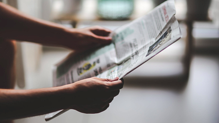 A person holds a newspaper open with outstretched arms.