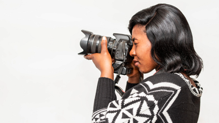 A black female photographer holds up a camera to her eye to take a photo (By Charles Wundengba from Pexels).