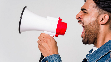 A person wearing a blue denim jacket yells into a red and white megaphone (By Sora Shimazaki from Pexels).