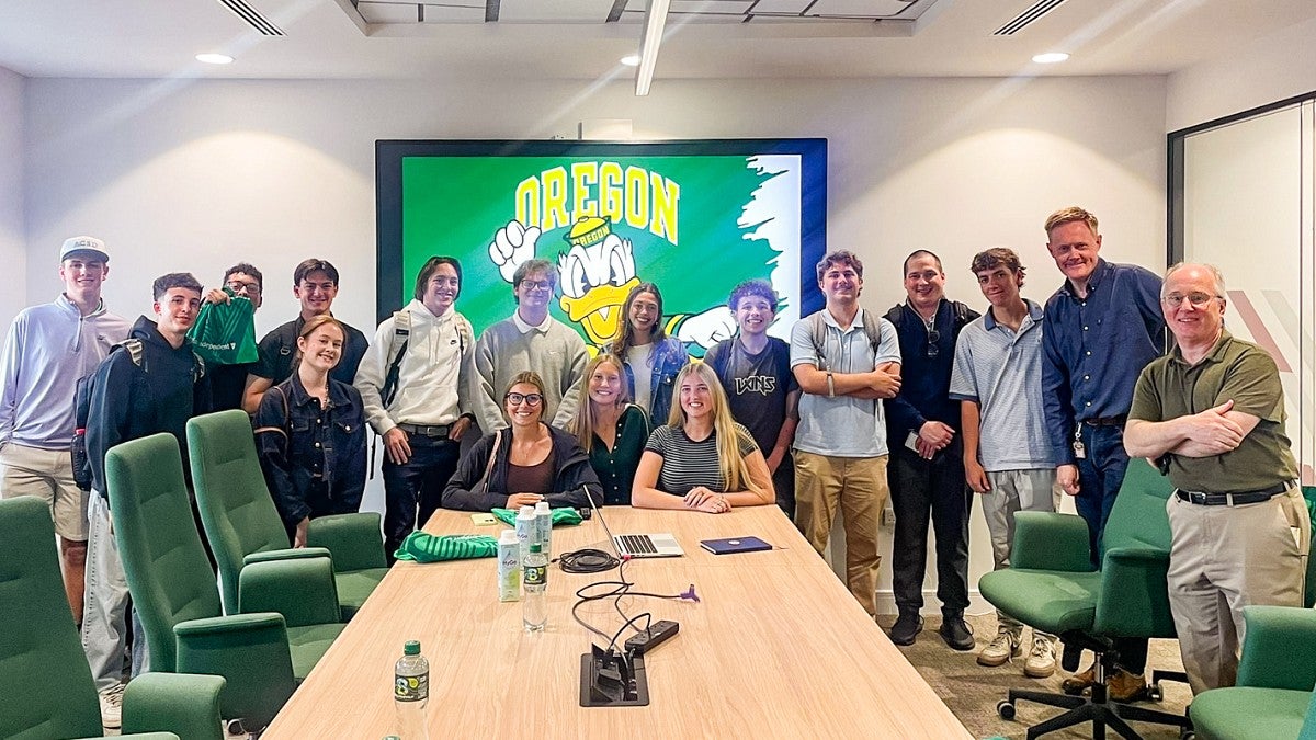 a group of University of Oregon School of Journalism and Communication students throw their Os as they pose in a conference room in front of a digital screen featuring the Oregon Duck 
