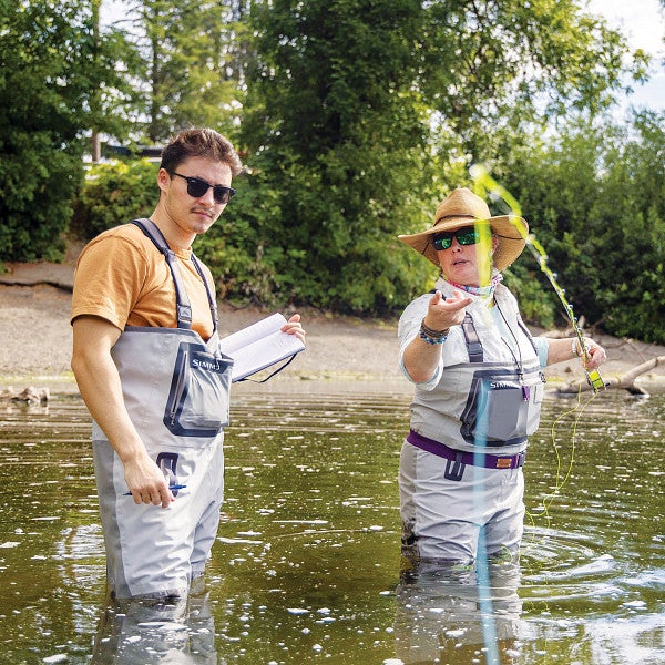 A student reporter interviews a person fly fishing while they both stand in a river