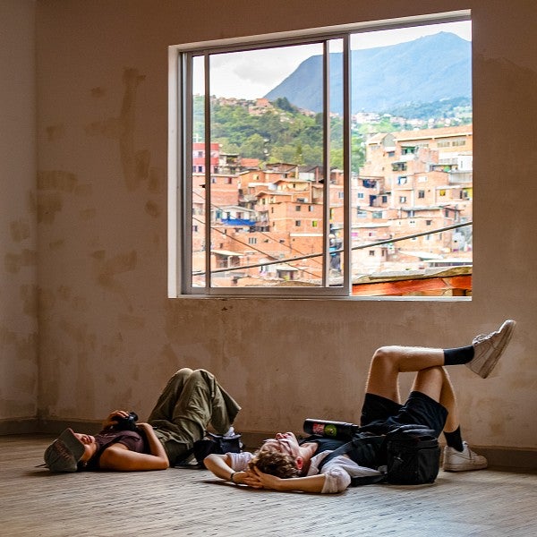 two students lie on the floor below an open window showing the hills of a city in Colombia