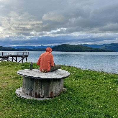 A person wearing an orange jacket sits on a wooden table outside in Cordova, Alaska