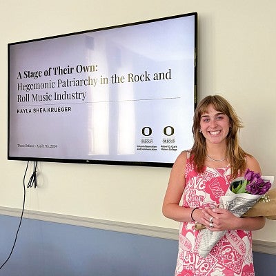 Kayla Krueger poses with flowers in front of a large screen showing her thesis title