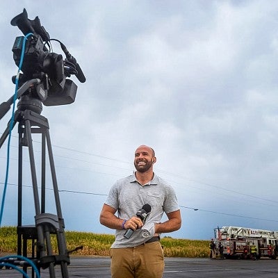 low angle shot of Zach Bruhl reporting TV news live from the field while standing in front of a video camera