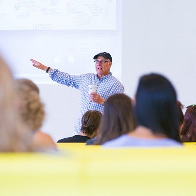 Dave Koranda gestures at a large display screen with the backs of students' heads in the foreground