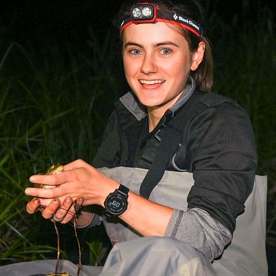 Eden McCall, wearing a headlamp and crouching in aquatic grasses at night, holds a small spotted frog, the subject of her honors thesis