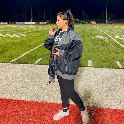 Sarah Ortiz speaks into a headset microphone from the sidelines of a football field