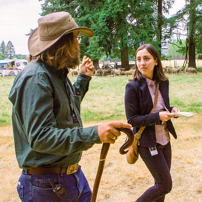 Francesca Fontana interviews a person who is facing away from a camera. They are outdoors in Oregon, with sheep in a pasture behind them. 
