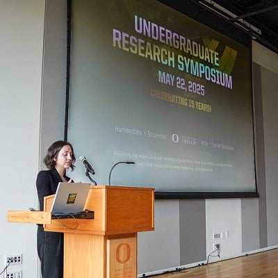 Francesca Fontana stands at a lectern in front of a large welcome screen at the 2025 UO Undergraduate Research Symposium keynote address