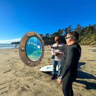 digital composite showing a person standing on the beach wearing XR goggles and reaching for a digital porthole into the ocean while another person watches